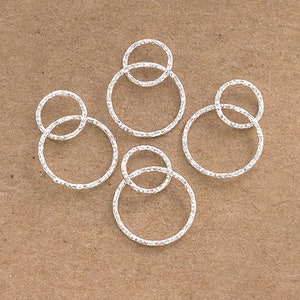 Pode incluir: Cinco anéis de prata, cada um com uma superfície texturizada, estão dispostos sobre um fundo castanho. Os anéis são de tamanhos diferentes e estão empilhados uns sobre os outros.