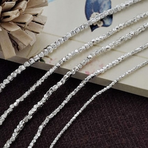 May include: Five strands of silver-coloured metallic beads arranged in parallel. Each strand consists of small, faceted, reflective beads strung together in a continuous line, showcasing varying thicknesses across the five individual chains.