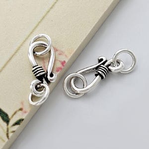 Op de afbeelding: Twee zilverkleurige haaksluitingen. Elke sluiting heeft een gebogen haak, een lusontwerp en een kleine ring. De sluitingen hebben een decoratief draadomwikkeld detail bij de haak. Ze worden waarschijnlijk gebruikt voor het maken van sieraden of soortgelijke ambachten.