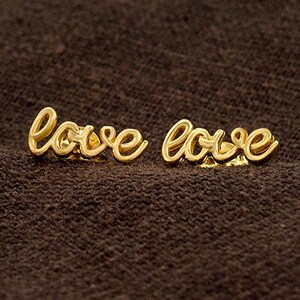 May include: A pair of gold-coloured earrings spelling out the word "love" in a cursive script. The earrings are displayed on a dark brown, textured surface.