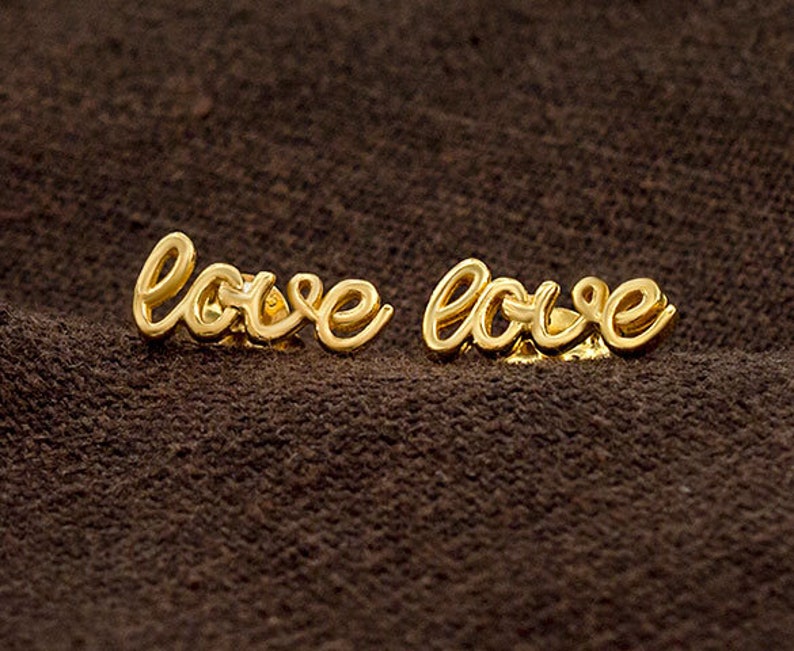 May include: A pair of gold-coloured earrings spelling out the word "love" in a cursive script. The earrings are displayed on a dark brown, textured surface.