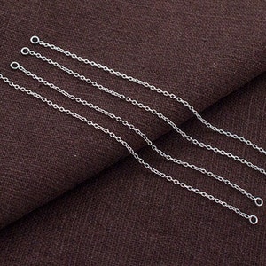 May include: Four delicate silver chain extenders with small circular loops at each end. The chains are laid out on a brown fabric surface, highlighting their fine links, suitable for jewellery making or repair.