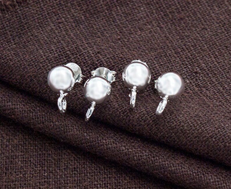 May include: Four silver earring backs with a round ball shape. The earring backs are on a brown fabric surface.