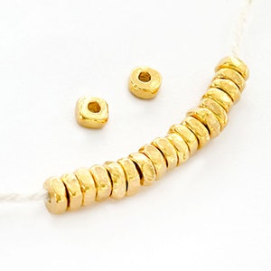 Puede incluir: Una hebra de cuentas de color dorado con textura martillada. Las cuentas están enhebradas en un cordón blanco y hay dos cuentas adicionales a un lado.