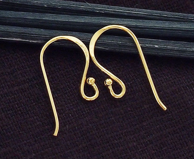 Op de afbeelding: Een paar goudkleurige metalen oorhaken met een gebogen draadontwerp en een kleine bolvormige kraal aan de basis van elke lus. Deze sieradenonderdelen zijn ontworpen voor het bevestigen van decoratieve elementen om afgewerkte hangende oorbellen te maken.
