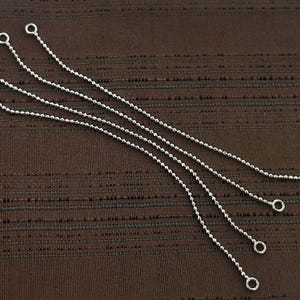 Pode incluir: Quatro puxadores de corrente de bolinhas prateadas com pequenos laços em cada extremidade, dispostos diagonalmente sobre um fundo castanho texturizado. Estes puxadores são provavelmente para luminárias ou aplicações semelhantes.