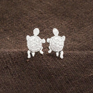May include: A pair of silver turtle stud earrings. The turtles are facing forward with their heads up and their legs outstretched.