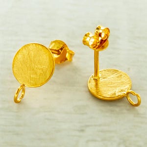 May include: Two gold-colored earring components. Each component has a round, flat disc with a textured surface and a small loop at the bottom. One component has a post and butterfly backing. The other component is missing the backing.