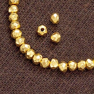 May include: A close-up shot of a string of gold-coloured faceted beads, with a few loose beads scattered around. The beads have a textured, irregular surface and are strung on a thin wire. The background is a dark brown fabric.