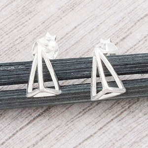 May include: A pair of silver-colored geometric stud earrings. Each earring features a triangular open frame design. The earrings are displayed on a textured, light-colored surface.