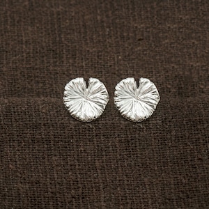 May include: A pair of silver earrings shaped like water lily leaves. The earrings are on a brown fabric background.