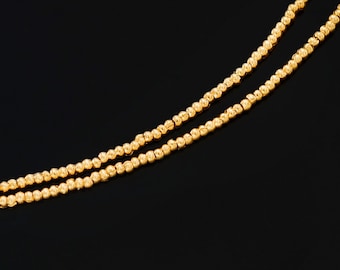 May include: Two strands of a gold-colored beaded necklace against a black background. The beads are small and closely spaced, creating a textured appearance. The necklace is angled across the frame.