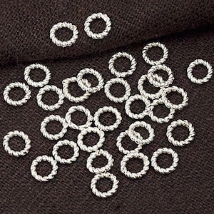 May include: A collection of silver jump rings, each with a twisted rope design, scattered on a brown fabric surface.