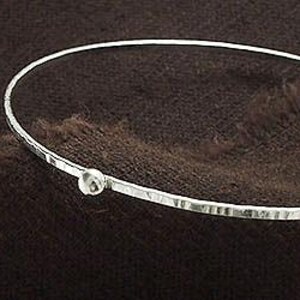 Op de afbeelding: Een zilverkleurige armband met een gehamerde textuur en een kleine, ronde, heldere steen. De armband is geplaatst tegen een donkerbruine, getextureerde achtergrond.