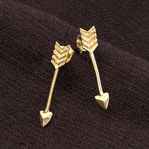 Könnte beinhalten: Ein Paar goldene Pfeilohrringe. Die Pfeile sind mit einem strukturierten Design verziert und haben eine spitze Spitze.