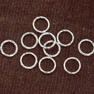 May include: A collection of small, silver-coloured rings with a textured, twisted design. The rings are scattered on a brown, textured surface. These rings are suitable for jewellery making and crafting. Each ring has a diameter of approximately 1 cm.