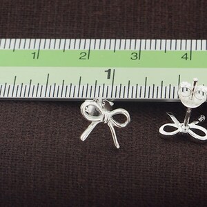 May include: A pair of silver bow-shaped stud earrings. The earrings are approximately 1 inch in length and are shown next to a ruler for size reference.