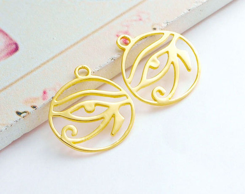 Puede incluir: Dos abalorios de metal de color dorado en forma de ojo con detalles intrincados. Los abalorios tienen un bucle en la parte superior para sujetarlos a joyas u otras manualidades.