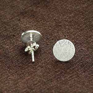 Könnte beinhalten: Zwei silberne Ohrstecker mit gebürstetem, kreisförmigem Design. Einer der Ohrringe zeigt den Stift und den Butterfly-Verschluss. Die Ohrringe werden auf einer braunen, strukturierten Oberfläche präsentiert.