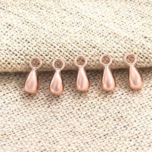 May include: Six rose gold teardrop shaped charms with a loop at the top. The charms are on a light brown fabric background.