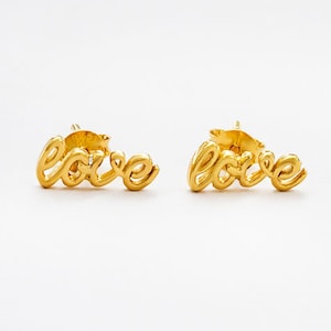 May include: A pair of gold-coloured stud earrings. Each earring spells out the word "love" in a cursive script. The earrings are set against a white background.