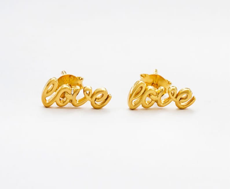 May include: A pair of gold-coloured stud earrings. Each earring spells out the word "love" in a cursive script. The earrings are set against a white background.