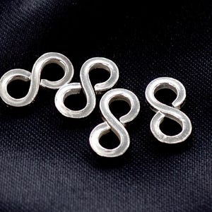 Puede incluir: Cuatro colgantes plateados con forma de símbolo de infinito. Los colgantes están dispuestos sobre una superficie oscura y texturizada. Cada colgante es un bucle continuo, con un diseño suave y redondeado. Estos colgantes podrían usarse para hacer joyas u otras manualidades.