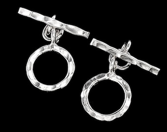 Può includere: Due chiusure a T in argento, ciascuna composta da un anello circolare e una barra dritta. Entrambi i pezzi presentano una superficie martellata e testurizzata che riflette la luce, creando un aspetto metallico brillante su uno sfondo scuro.