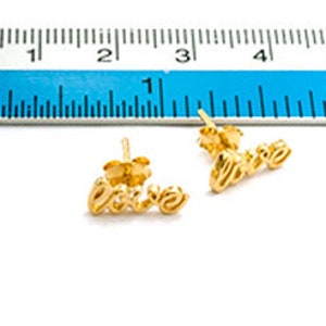 May include: A pair of gold-coloured stud earrings spelling out the word "love" in cursive script. The earrings are approximately 2 cm long, with a post backing. A blue ruler is in the background.