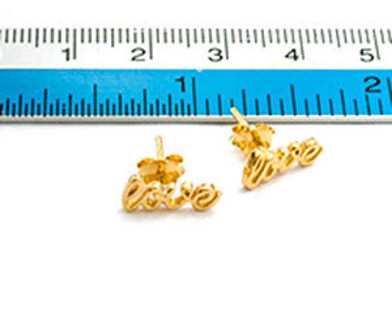 May include: A pair of gold-coloured stud earrings spelling out the word "love" in cursive script. The earrings are approximately 2 cm long, with a post backing. A blue ruler is in the background.