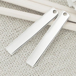 May include: Two silver metal bar pendants with a hole at each end. The pendants are laying on a white fabric surface.