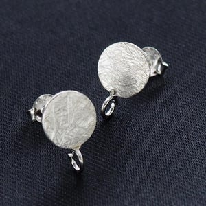 Pode incluir: Um par de brincos de prata. Cada brinco apresenta um disco circular texturizado com um pequeno laço e um pino. Os brincos estão sobre um fundo escuro, realçando o seu brilho metálico. Joalharia.