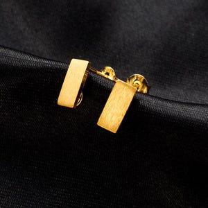 Puede incluir: Un par de pendientes de botón rectangulares de color dorado. Los pendientes tienen un acabado cepillado.