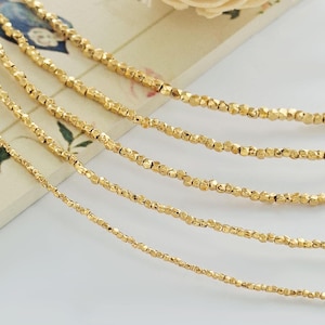 May include: A collection of five gold-toned chains, each featuring a series of small, faceted, irregular beads strung together. The chains are arranged in a cascading, layered formation, showcasing the reflective, textured surface of the metallic beads.