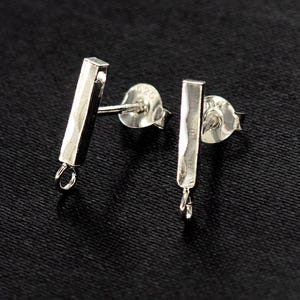 May include: A pair of silver bar stud earrings with a textured finish. Each earring has a post for piercing and a small loop at the bottom. The earrings are displayed on a black surface.
