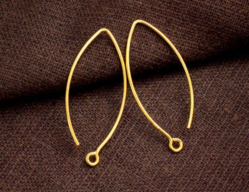 May include: Two gold-toned earring hooks, each with a slightly oval shape, are shown against a brown fabric background.