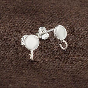 Könnte beinhalten: Ein Paar silberne Ohrring-Verschlüsse mit einer runden Scheibe und einer kleinen Schlaufe. Die Verschlüsse befinden sich auf einer braunen Oberfläche.