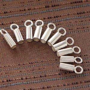 Peut inclure: Plusieurs embouts à sertir argentés avec de petites boucles. Ces éléments de bijouterie cylindriques sont conçus pour être fixés aux chaînes ou à d'autres composants. Les embouts sont disposés sur une surface marron texturée.