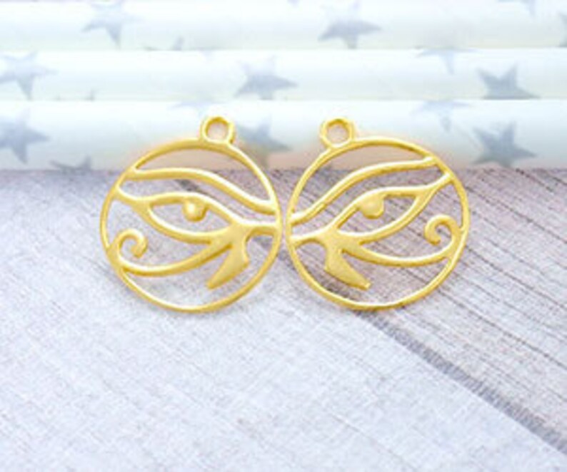 Puede incluir: Dos abalorios de metal de color dorado en forma de c&iacute;rculo con un dise&ntilde;o de ojo. Los ojos son estilizados y tienen un patr&oacute;n de espiral alrededor de ellos.