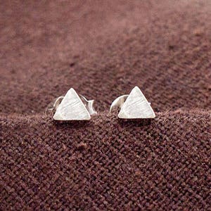 Peut inclure: Une paire de petites boucles d'oreilles triangulaires argentées. Les boucles d'oreilles ont une surface texturée et sont posées sur un fond marron texturé. Les boucles d'oreilles sont de conception géométrique simple.