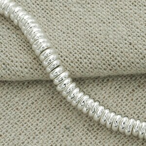 Puede incluir: Un brazalete de cadena de plata hecho de peque&ntilde;os eslabones redondos conectados. La cadena est&aacute; sobre una superficie de tela beige texturizada.