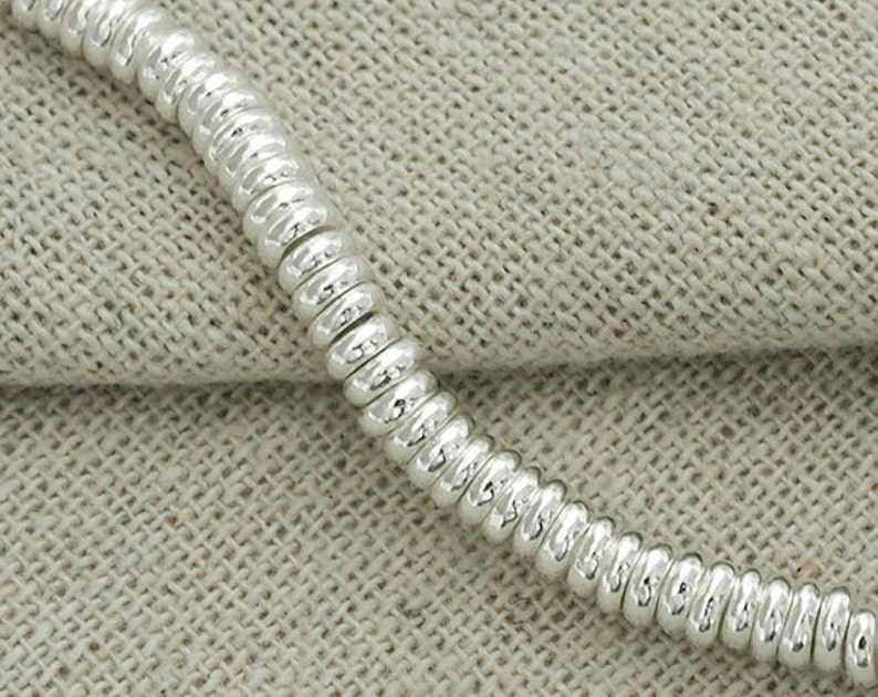 Puede incluir: Un brazalete de cadena de plata hecho de peque&ntilde;os eslabones redondos conectados. La cadena est&aacute; sobre una superficie de tela beige texturizada.