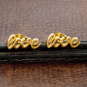 May include: A pair of gold-coloured stud earrings. Each earring spells out the word "love" in a cursive font. The earrings are displayed on a dark, textured surface against a brown background.