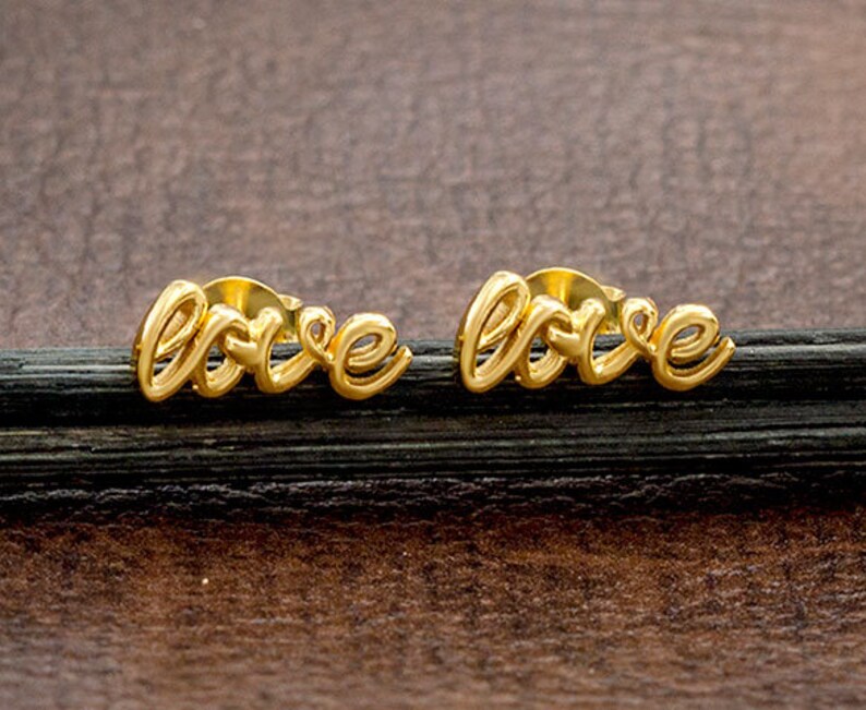 May include: A pair of gold-coloured stud earrings. Each earring spells out the word "love" in a cursive font. The earrings are displayed on a dark, textured surface against a brown background.