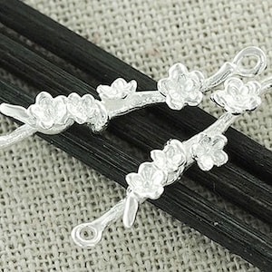 Puede incluir: Dos colgantes plateados con forma de rama floral. Cada colgante presenta una rama con delicadas flores y un lazo en cada extremo. Los colgantes se muestran sobre una superficie texturizada de tonos neutros con elementos lineales oscuros.