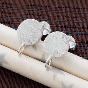 May include: A pair of silver-colored stud earrings with a circular design and a textured surface. The earrings have a post and a loop for attaching to the ear. The earrings are displayed on a white and gray star-patterned paper.