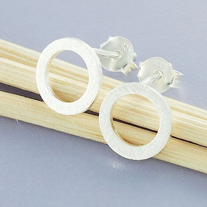 Könnte beinhalten: Ein Paar silberfarbene Ohrstecker mit offenem Kreis. Die Ohrringe haben eine strukturierte Oberfläche und werden auf einer hellen Holzoberfläche vor einem weichen grauen Hintergrund präsentiert. Die Ohrringpfosten und -verschlüsse sind sichtbar.