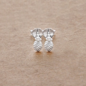 May include: A pair of silver pineapple stud earrings. The earrings are small and delicate, and they feature a realistic pineapple design.