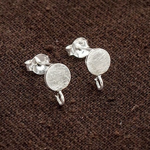 Könnte beinhalten: Zwei silberne Ohrstecker mit kreisförmigem Design und strukturierter Oberfläche. Jeder Ohrring hat einen Stift zum Einsetzen und eine kleine Schlaufe zum Anbringen von Anhängern. Präsentiert auf einem braunen Stoff.