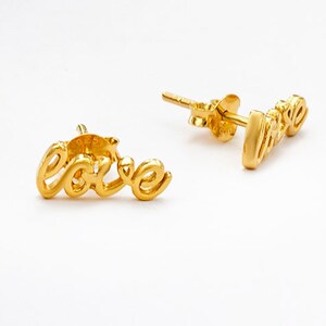 May include: A pair of gold-coloured stud earrings. Each earring is shaped to spell out the word "love" in a cursive font. The earrings are set against a white background.